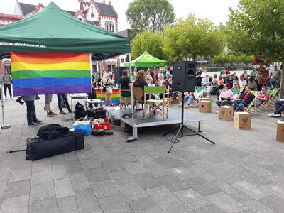 Foto der Bühne von hinten mit Regenbogenfahne. Zahlreiche Zuschauer haben sich versammelt und diskutieren mit.